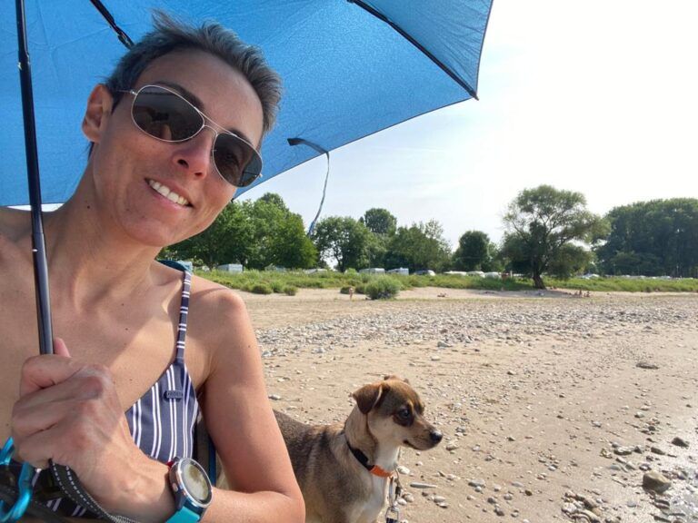 Lucie Petrelis life coaching for multiple sclerosis relaxing at the beach with her small size dog while holding a small size foldable umbrella to protect herself from the strong sun.